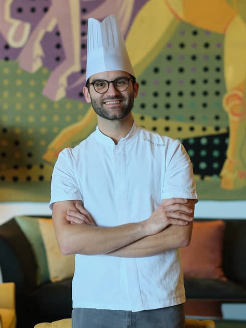 A chef in a white shirt and toque stands with arms crossed in a cozy interior space with a table lamp, upholstered chair and couch, and a mural on the back wall