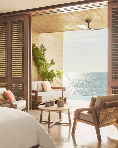 A serene bedroom with wooden accents, featuring a seating area that opens to a terrace overlooking the ocean. Soft natural light fills the room, highlighting the warm, earthy decor.