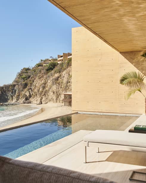A luxurious oceanfront terrace with an infinity pool, lounge chairs and outdoor dining, overlooking a secluded beach and rocky cliffs under a bright blue sky