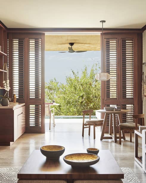 Bright living area with wooden shutters opening to an outdoor terrace. The room features a grey sofa, a small dining table and elegant wooden furniture, with a view of greenery outside.
