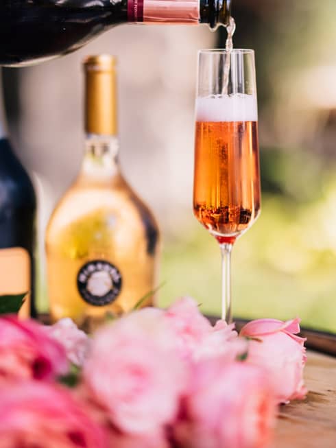 A glass of rose next to rose petals and bottles.