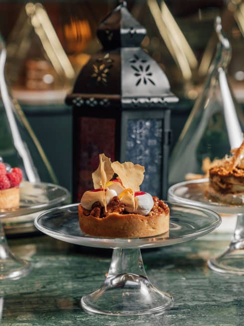 Three desserts each plated on their own glass cake stand are set next to each other on a blue-green granite table with a traditional Arabian-style lantern behind and center