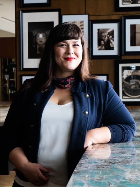 A female staff member in a blue jacket, smiling behind a bar of a restaurant with dim lighting.