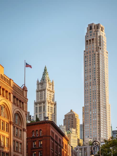 Downtown cityscape view featuring historic and modern skyscrapers, including the Woolworth Building, with an American flag against a clear blue sky
