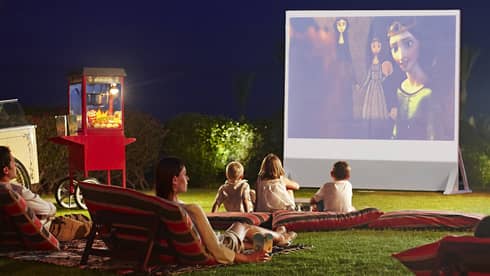 Children, parents lounge on pillows on grass in front of large outdoor movie screen at night