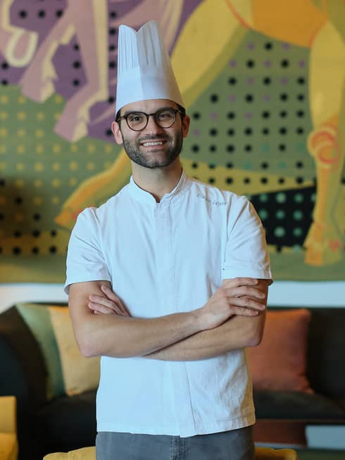 A chef in a white shirt and toque stands with arms crossed in a cozy interior space with a table lamp, upholstered chair and couch, and a mural on the back wall