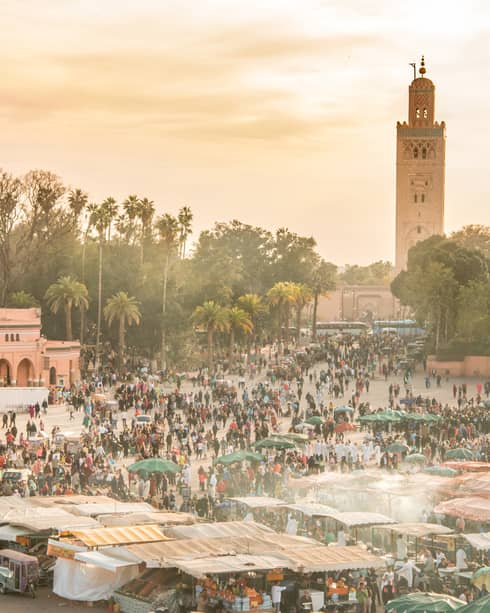 Hazy overhead view of an outdoor market teeming with people, stalls and umbrellas, with trees and a tall minaret at the back.