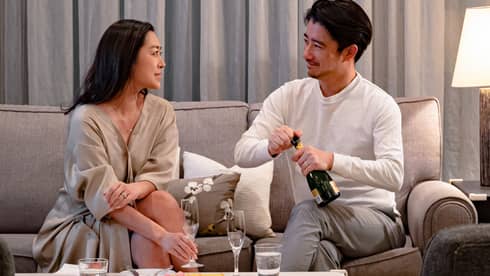 Two people sitting on a sofa, smiling as one opens a bottle of champagne