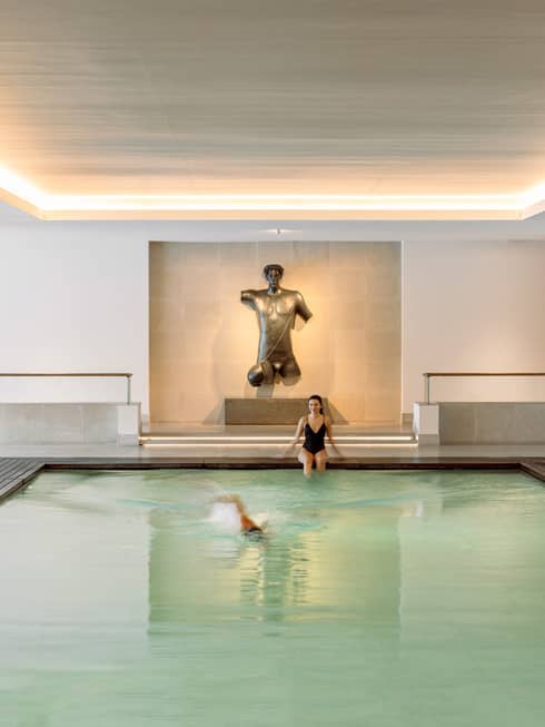 Person sitting at end of indoor lap pool under uplit ceiling while another person swims