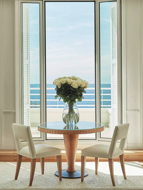 White chairs by small cafe table, large glass vase with flowers, sunny window