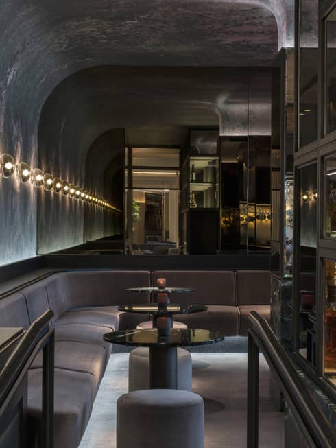 A velvet banquet and three round cocktail tables line the curved wall of MARCUS bar next to glass shelves holding bottle of spirits, all dimly lit