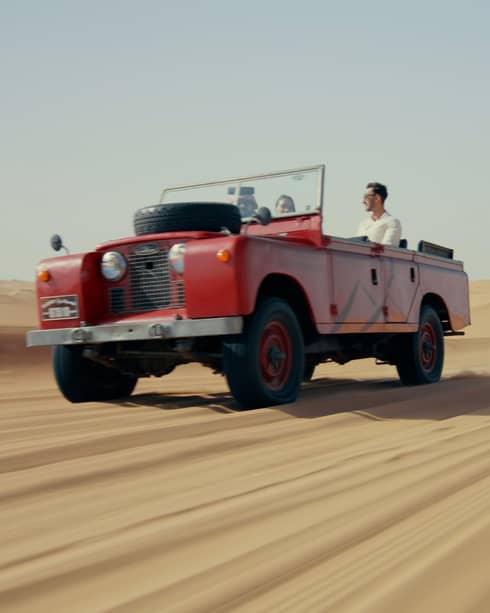 A red truck driving down a long stretch of sand under a blue sky.