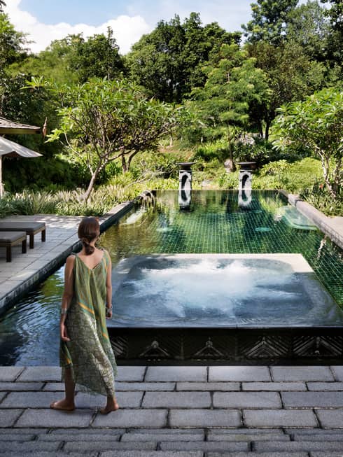 Back of woman in long beach dress walking towards residence swimming pool surrounded by tropical forest
