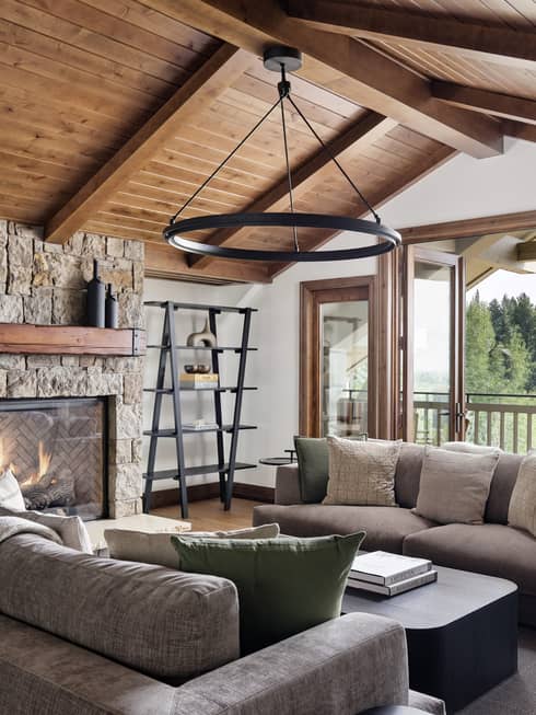 Granite residence living room with vaulted wood ceiling, stone fireplace, grey sofas and a circular chandelier, opening to a balcony with forest view