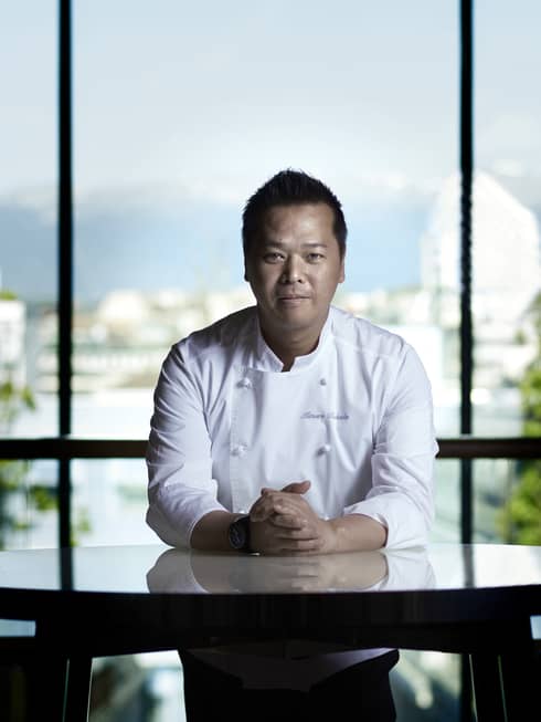 Portrait photo of Chef de Cuisine Mitusuru Tsukada in white chef's coat, at table in front of sunny window