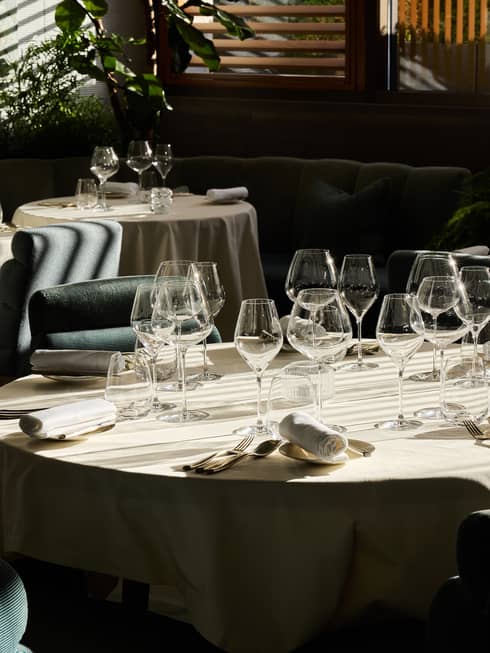 An indoor dining space with dramatic daylight filtering through window blinds and featuring tables, chairs, and place settings for a meal