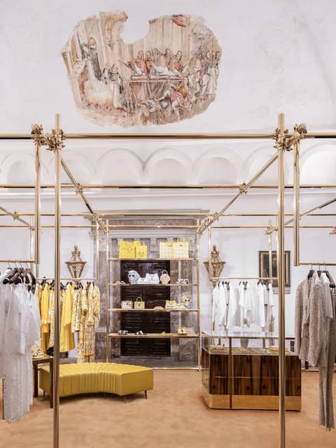 Elegant boutique with gold bars displaying white, tan and yellow clothes and suits under domed frescoed ceiling