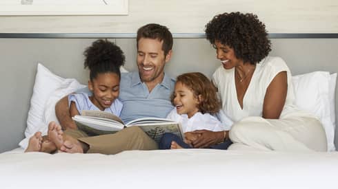 A happy family of four sits on a bed, smiling and reading a book together in a cozy, modern bedroom.