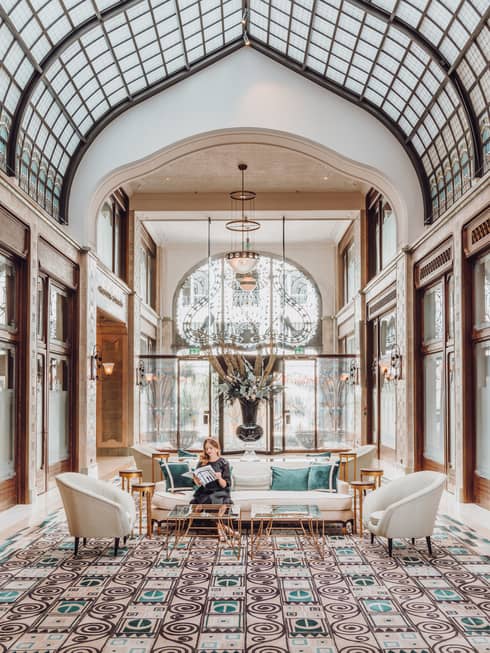 Elegant hotel lobby with arched glass ceiling, decorative floor tiles, plush seating and a large floral arrangement. A person sits on a sofa reading a magazine.