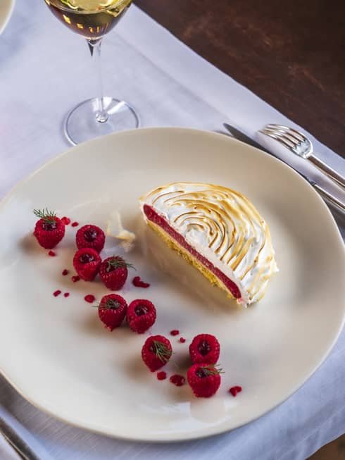 Half-circle cake topped with toasted meringue with line of raspberry garnish on round ecru plate