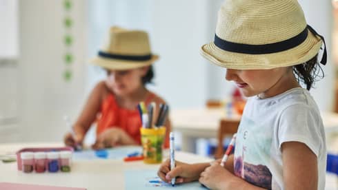 Two smiling children wearing straw fedora hats colour pictures with markers at craft table