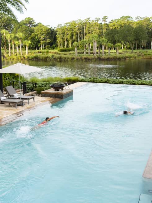 Two people swim in a large pool with two sets of steps leading into it. The pool is flanked by deck areas with lounge chairs, umbrellas, and palm trees and other plantings. A natural-looking body of water and other plants and trees are in the background.