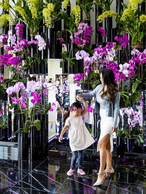 A woman and young girl surrounded by purple flowers and green plants.