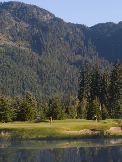 Distant golfers on a green, a pond in front, evergreens behind, dwarfed by a treed mountain filling most of the frame beyond.