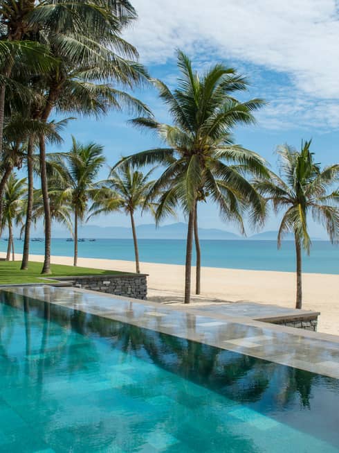 Tall palm trees between stone outdoor swimming pool and white sand beach