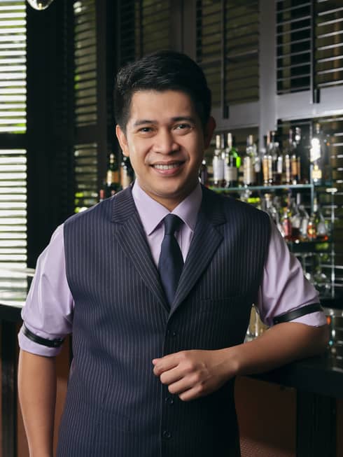 Smiling Bar Manager Gabriel Carlos poses for the camera, an artful display of liquor bottles on the shelf behind him