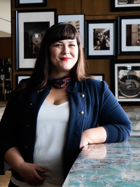 A female staff member in a blue jacket, smiling behind a bar of a restaurant with dim lighting.