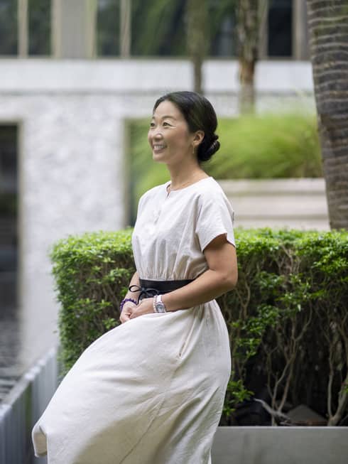 Smiling Asian woman in linen dress stands beside outdoor fountain