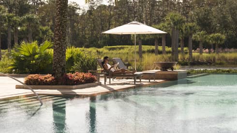 Outdoor swimming pool where woman lounges on patio chair under palm tree 