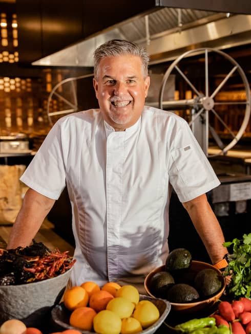 A smiling chef in white chefs clothes standing behind bowls of fruit and vegetables.