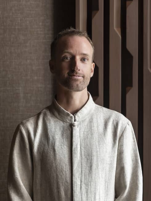 Smiling man with dark blonde mustache and beard in linen shirt stands in front of brown wall