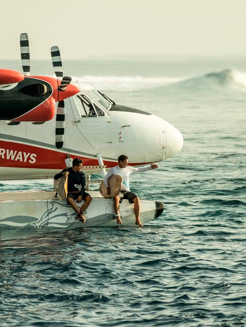 Two people holding surfboards sit on edge of float plane over water 