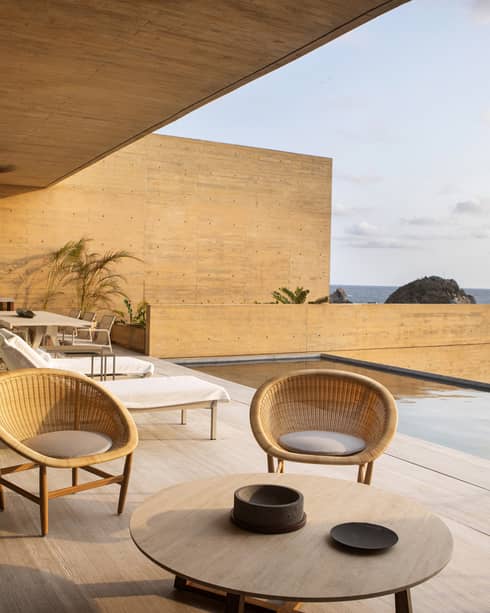 A modern oceanfront terrace featuring wicker chairs, a dining area and lounge chairs beside an infinity pool, all bathed in warm sunset light with a view of the sea
