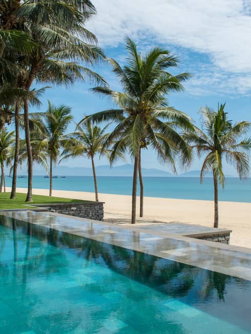 Tall palm trees between stone outdoor swimming pool and white sand beach