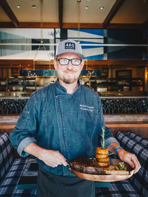 A smiling Chef Michael Goralski holds a skillet of steak and an onion ring tower