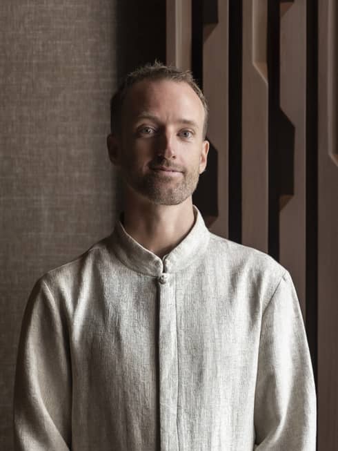 Smiling man with dark blonde mustache and beard in linen shirt stands in front of brown wall