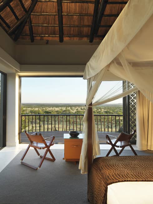 Poster bed, canopy by fold-up chairs, wood table by open wall with Serengeti views