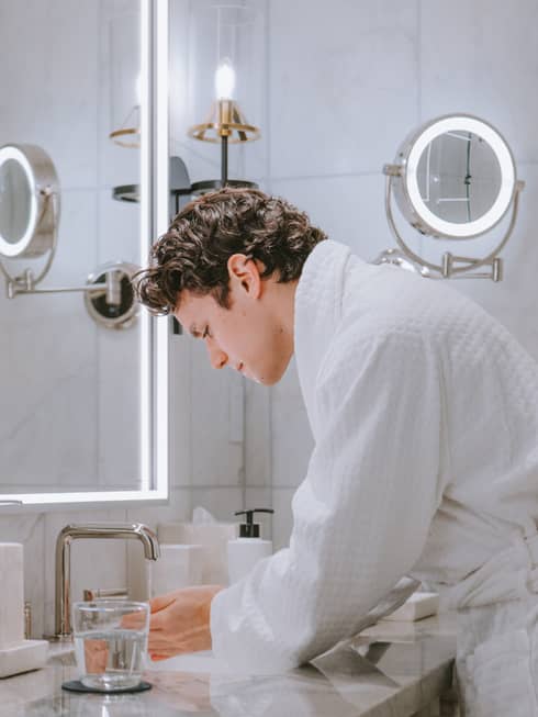 Person wearing a bath robe and washing their hands in the sink with a glass of water on the counter.
