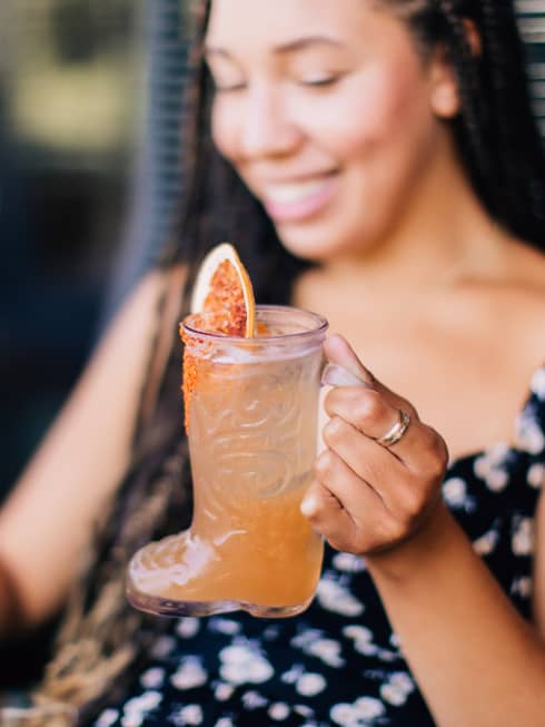 A woman in a black dress drinking an orange coloured cocktail.