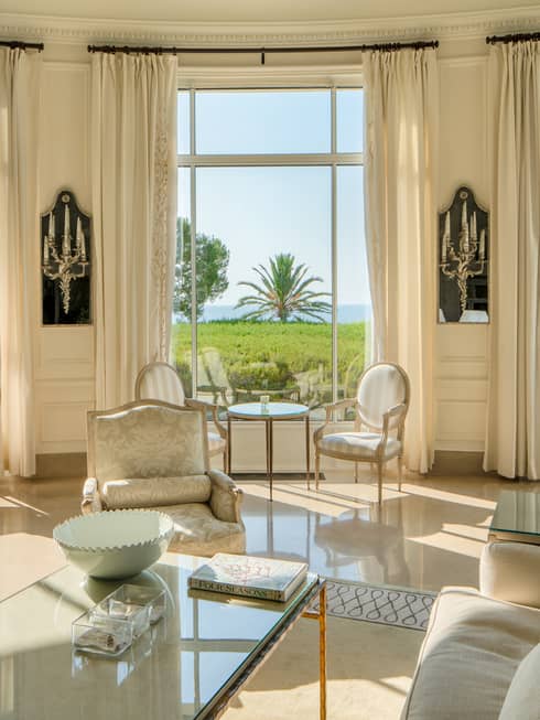All-white salon with couches and dining chairs with tall rounded windows, lawn and sea view