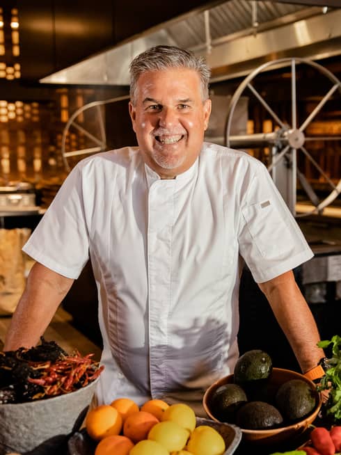 A chef smiling behind bowls of fruit and vegetables in a large kitchen.