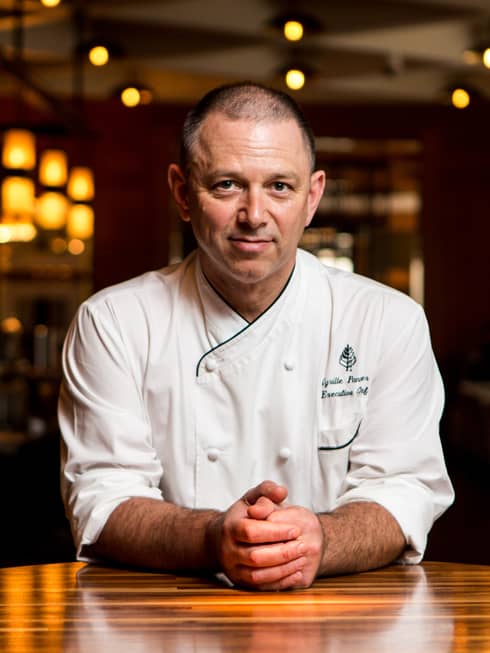 Portrait photo of Executive Chef Cyrille Pannier wearing white uniform, sitting at wood table with hands clasped in front