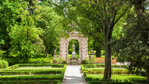 Garden with shrubs, trees in front of stone arch, statue