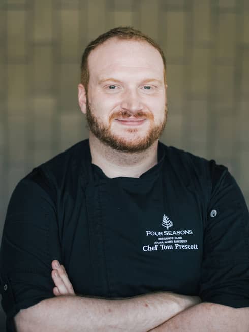 A male chef crossing his arms in black chef clothes in front of a brick wall.