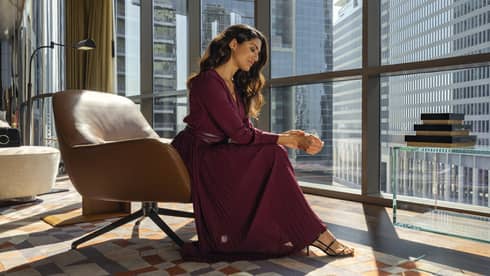 Woman in long burgundy dress sits next to floor-to-ceiling windows in sunny suite
