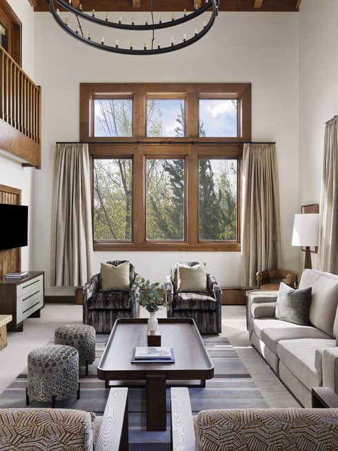 Spacious living room with stone fireplace, high ceiling, sofa, patterned armchairs, ottomans, tables, TV and tall windows overlooking trees